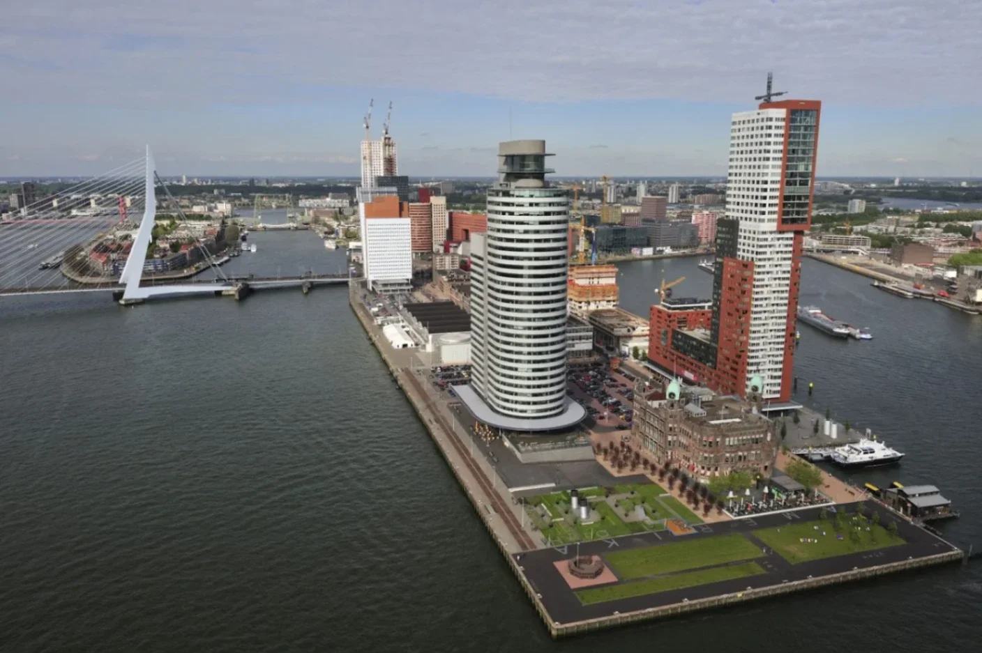 Modern cityscape featuring a mix of high-rise buildings along a waterfront, with boats docked and greenery in the foreground.