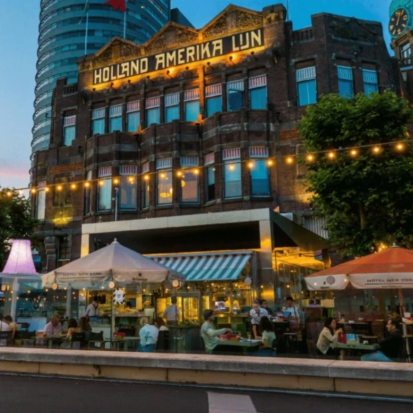 Historic building with "Holland Amerika Lin" signage, surrounded by lit outdoor dining areas and trees against a twilight...