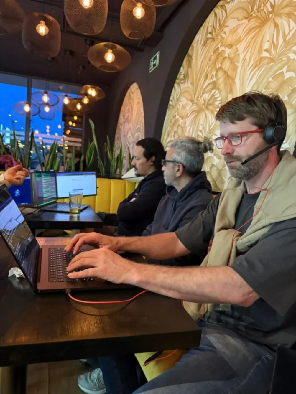 Three people work on laptops in a modern café, with Gconf's founder devving hard and focussed.